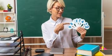 pleased young blonde female teacher wearing glasses sitting at desk with school tools in classroom showing and pointing at number fans looking at them